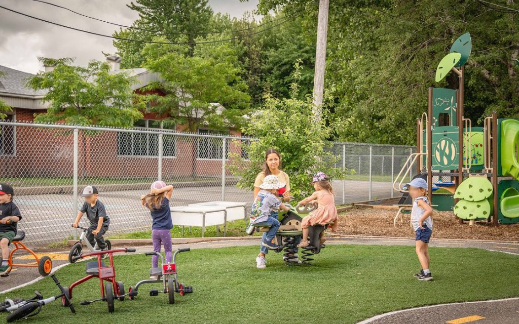 Cour extérieure, des enfants jouent sur un module de parc avec l'éducatrice et d'autres font du tricycle