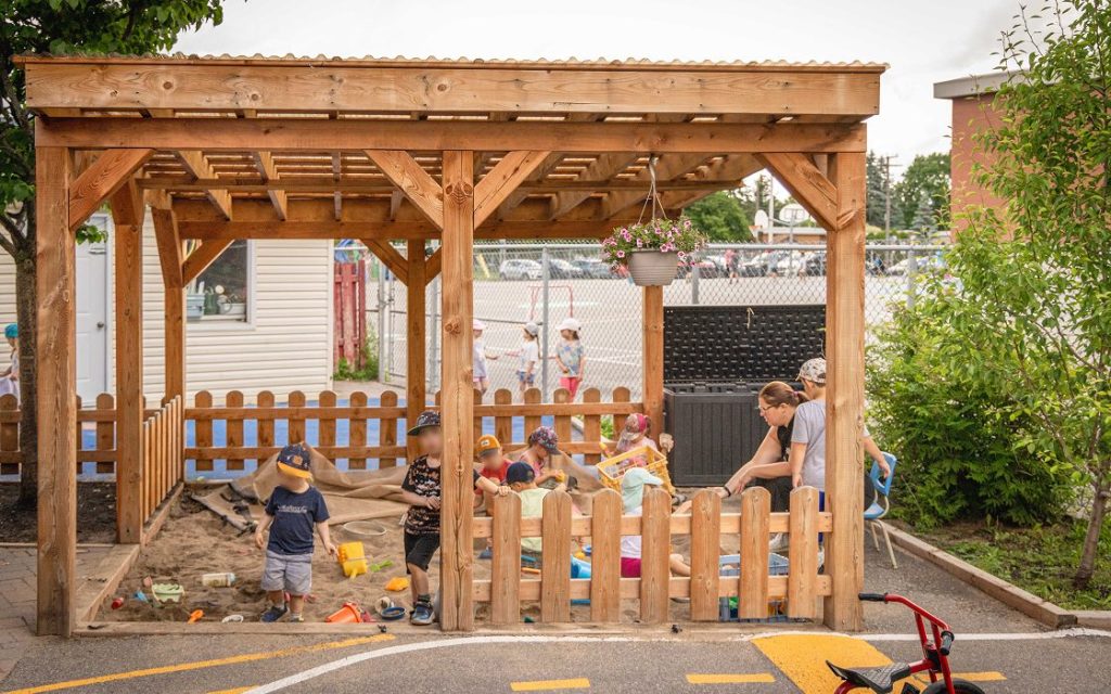 Une éducatrice joue avec les enfants dehors dans un grand bac à sable sous une pergola